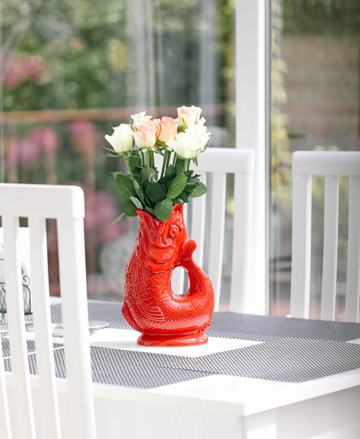 Eine leuchtend rote Gluckigluck Karaffe X-Large Rot von Gluckigluck, gefüllt mit weißen und hellrosa Rosen, steht auf einem modernen Esstisch mit einem grauen Tischset, umgeben von weißen Stühlen und großen Fenstern im Hintergrund.
