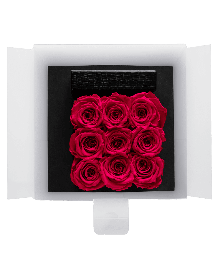 Die „Ivoryi Flowerbox Medium Black New Pink“ von Ivoryi & Friends enthält neun blühende rosa Rosen in einer weißen Box mit offenen Klappen auf schwarzem Untergrund, gekrönt von einem rechteckigen schwarzen strukturierten Akzent.