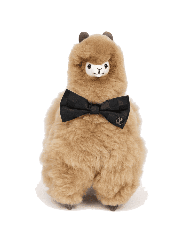 Das Alpaka-Stofftier PACO Champagne von Weich Couture Alpaka hat ein flauschiges hellbraunes Royal-Alpaka-Fell, kleine schwarze Ohren, ein weißes Gesicht und ist mit einer gemusterten schwarzen Fliege und einer Anstecknadel vor einem schlichten Hintergrund bekleidet.