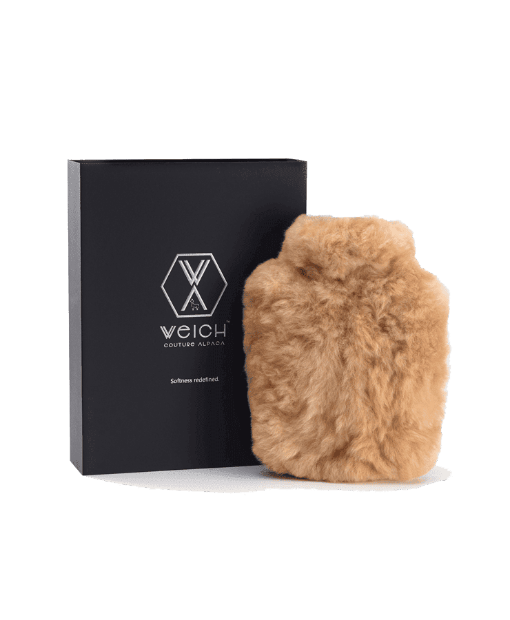 Eine flauschige, hellbraune Alpaka Wärmflasche Regular Champagne von Weich Couture Alpaka wird neben einer schwarzen Box mit dem Markenlogo und dem Satz „Softness redefined – Allergiker geeignet“ für ultimativen Komfort gezeigt.