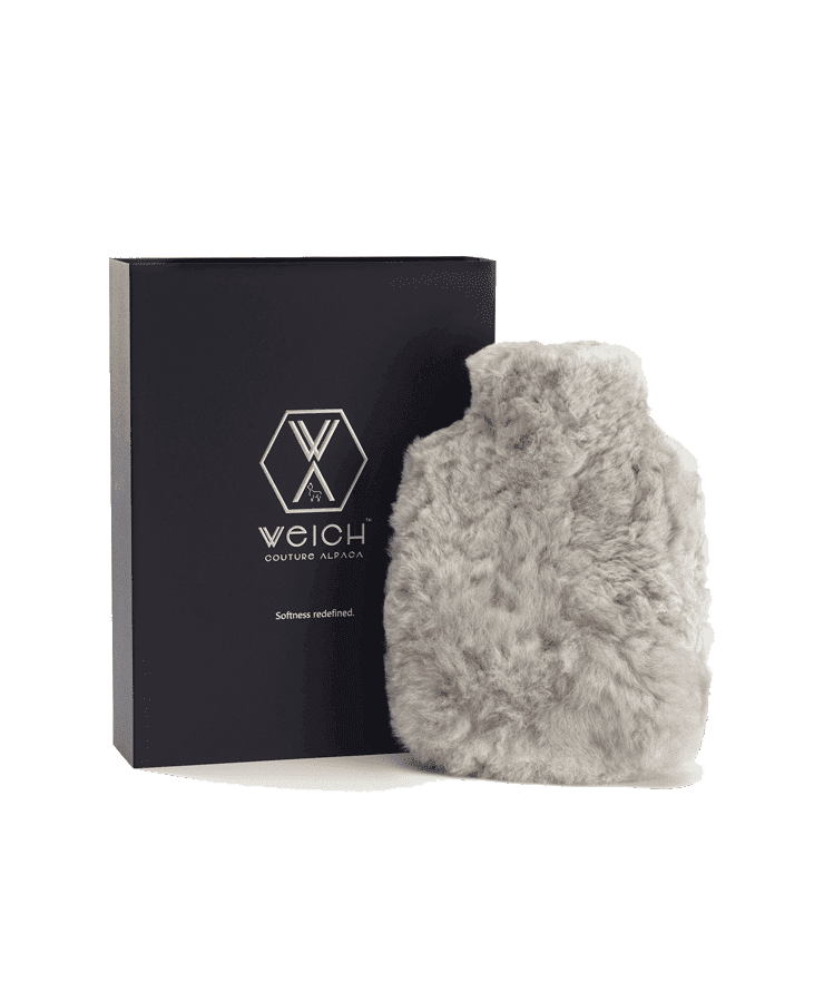 Die Alpaka Wärmflasche Regular Silver Grey von Weich Couture Alpaka mit einem weichen, pelzartigen silbergrauen Überzug wird neben einer schwarzen Box mit dem Markenlogo und „Softness redefined“ gezeigt, die an Hugo Froschs Royal Alpaca-Fell-Stil erinnert.