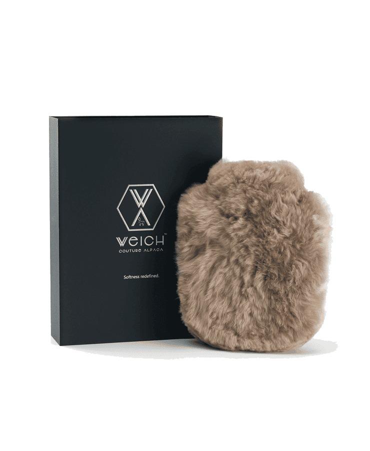 Die Alpaka Wärmflasche Regular Taupe von Weich Couture Alpaka ist ein plüschiger brauner Beutel aus Royal Alpaca-Fell, abgebildet neben einer eleganten schwarzen Box mit dem Markenlogo und „Softness redefined“ auf der Vorderseite. Für Allergiker geeignet.