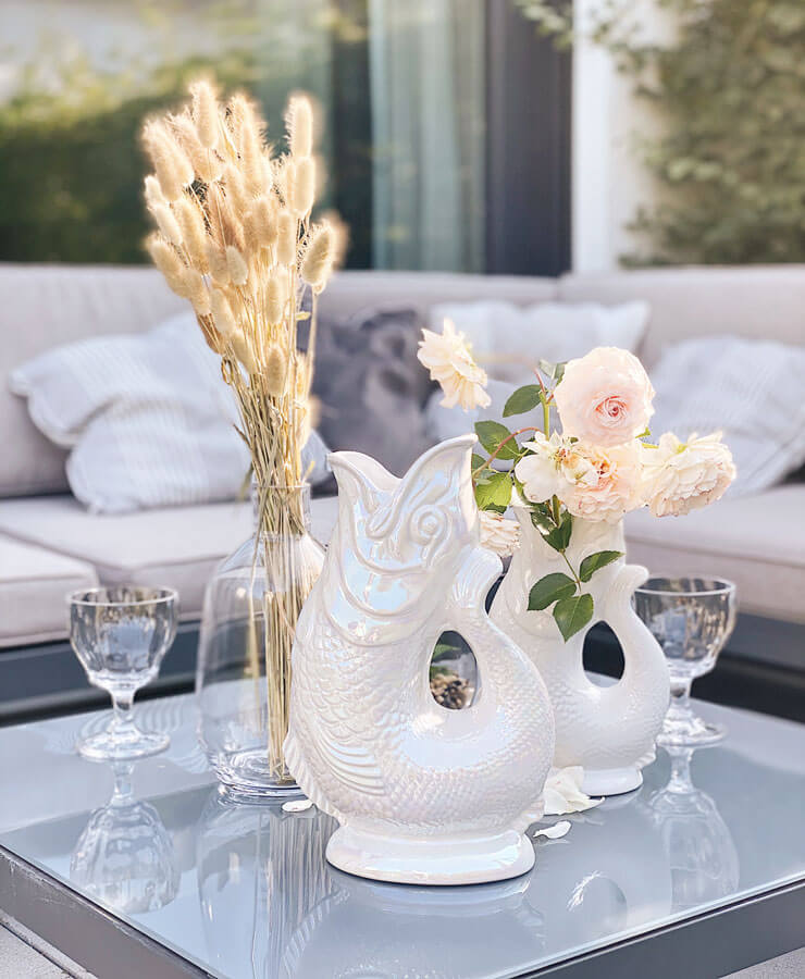 Ein moderner Couchtisch für den Außenbereich mit zwei Gluckigluck Karaffe X-Large Perlmutt von Gluckigluck, einer Glasvase mit getrockneten Gräsern, blassrosa Rosen und Kelchen – vor einem hellgrauen Sofa für entspannte englische Eleganz.